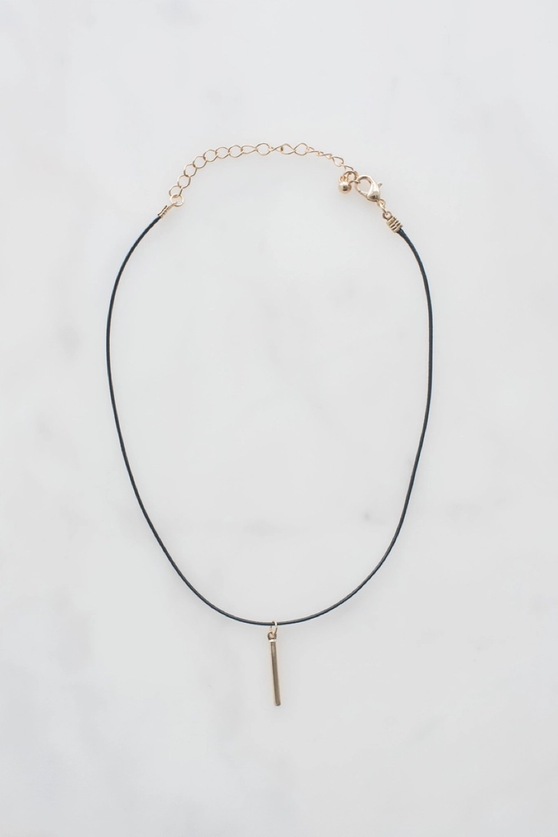A black cord choker with a gold bar pendant, laid flat on marble