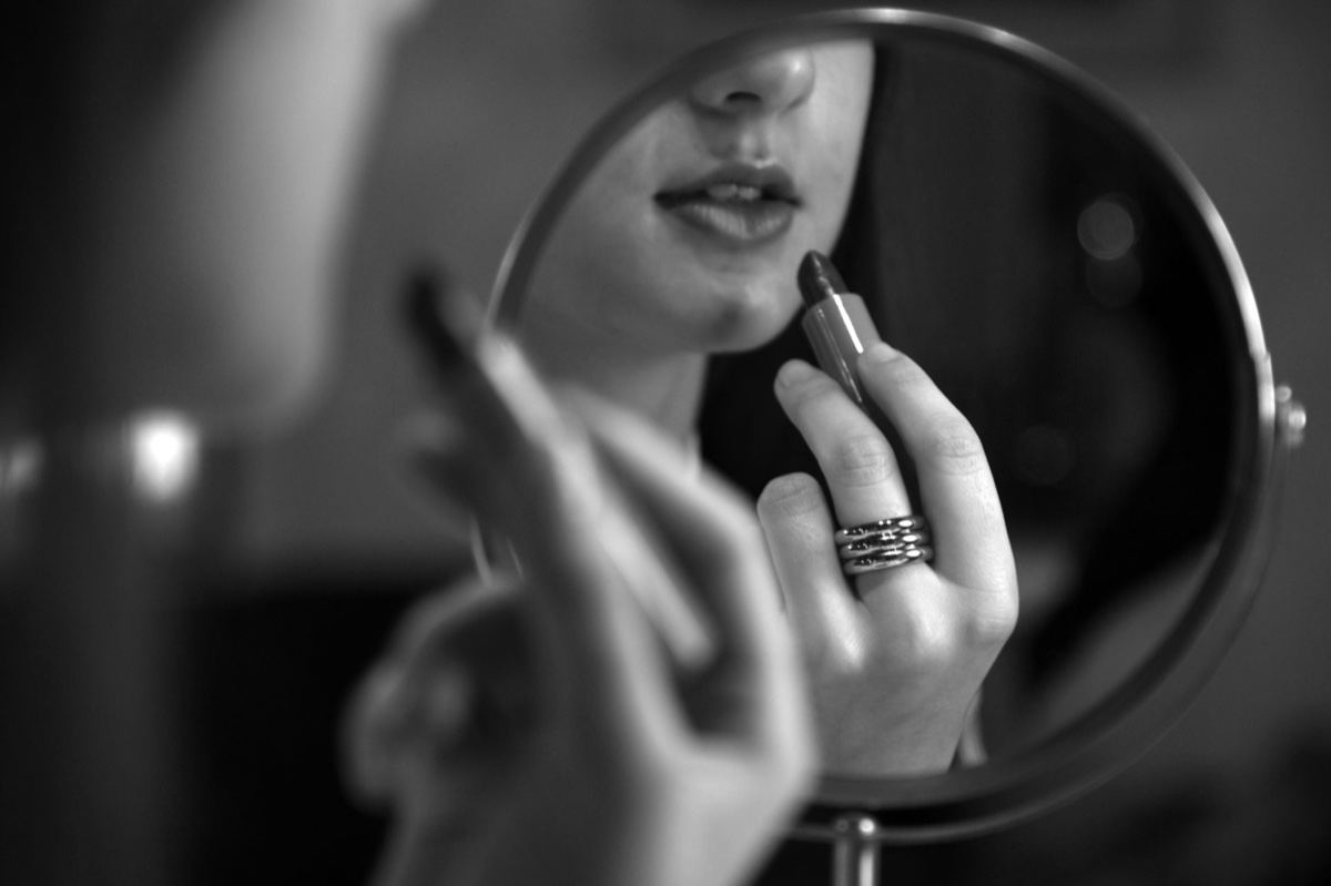 A woman applying dark lipstick, seen in the reflection of a round vanity mirror, in black and white