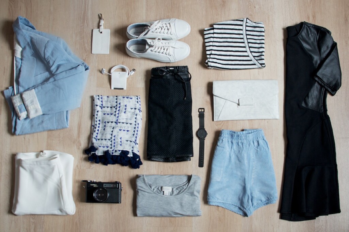 A small travel capsule laid out on a wood floor: shirts, shorts, sneakers, a clutch, a camera and a watch