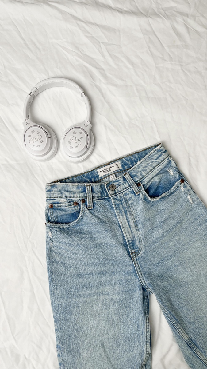 A pair of light-wash high-rise straight jeans laid flat on a white duvet next to a pair of white over-ear headphones
