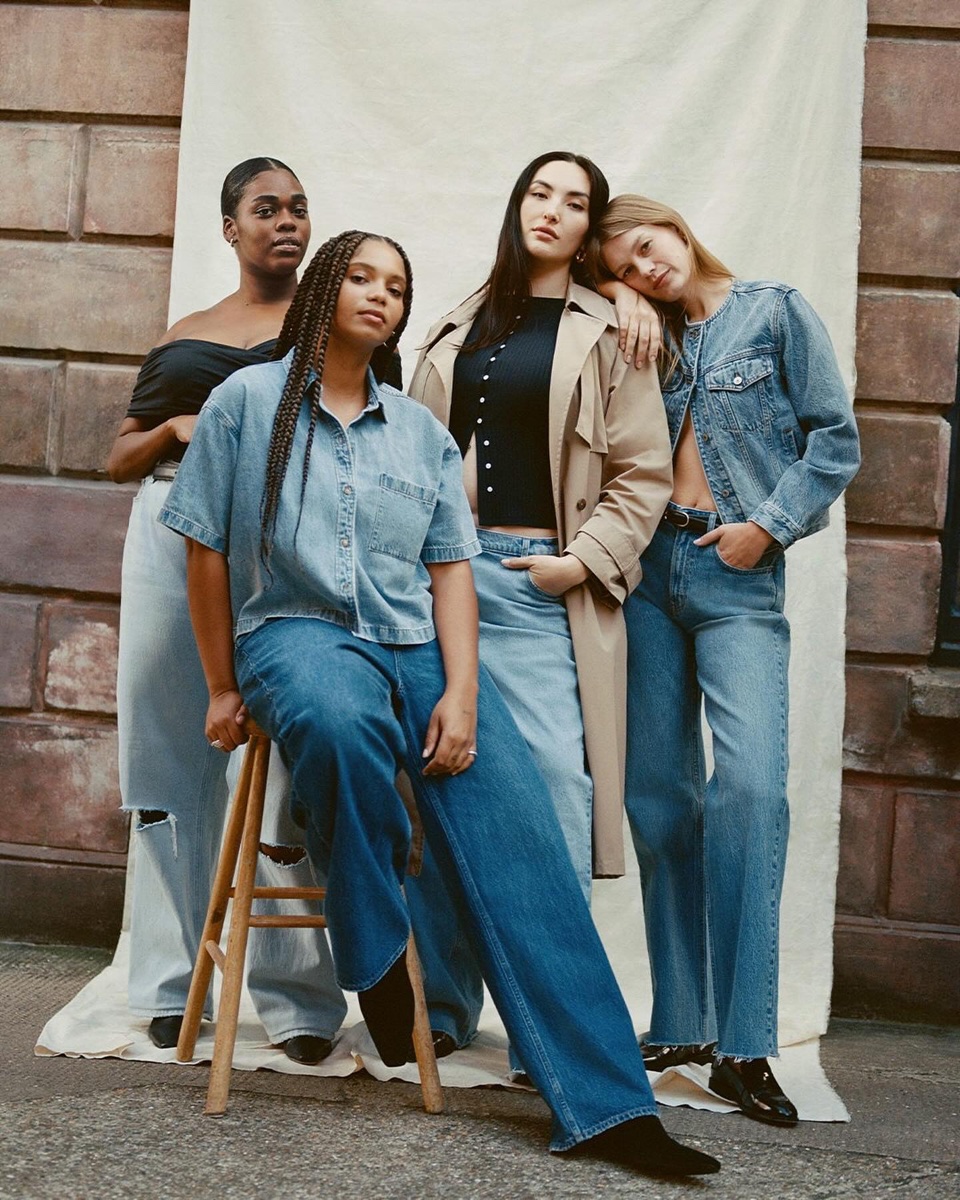 Four women in a range of blue denim, one seated on a wooden stool, posed in front of a draped canvas backdrop against a brick wall