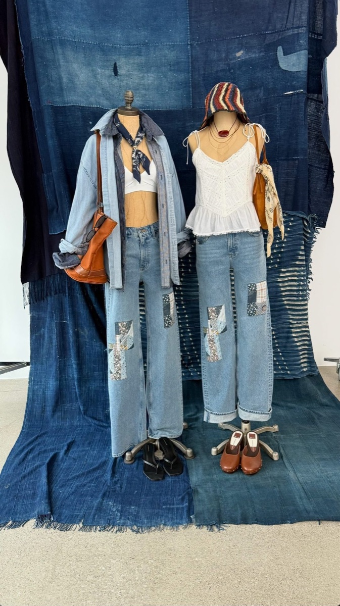 Two dress forms in light-wash patchwork jeans, one in a chambray shirt and one in a white eyelet camisole, against draped indigo cloth