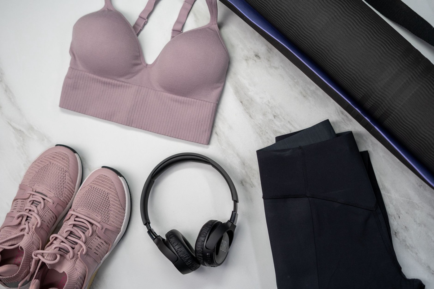 Flat lay of a pink sports bra, pink sneakers, black leggings, headphones and a yoga mat on marble