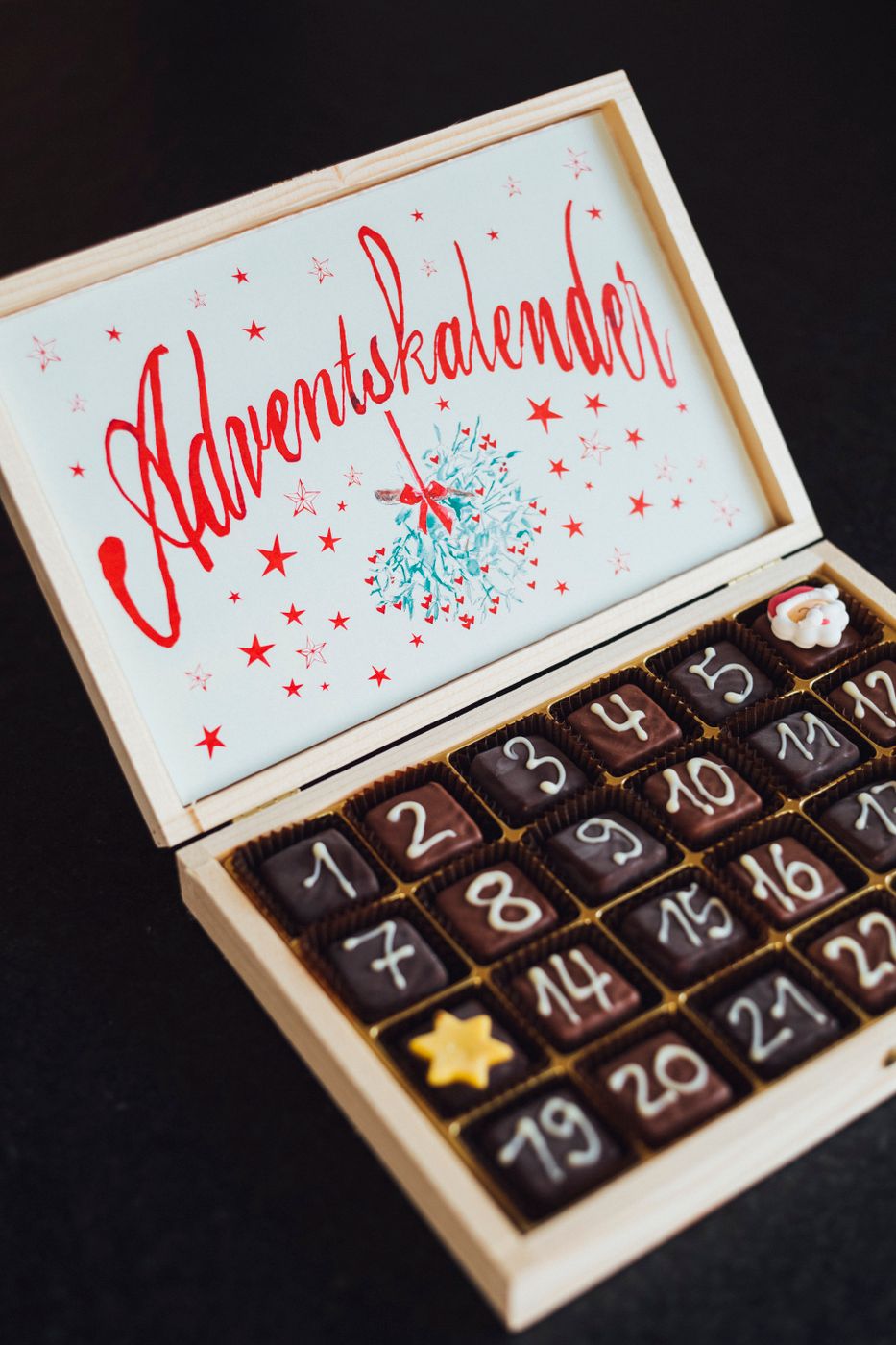 Open wooden advent calendar box with twenty-four numbered chocolates arranged in a grid under a hand-lettered lid