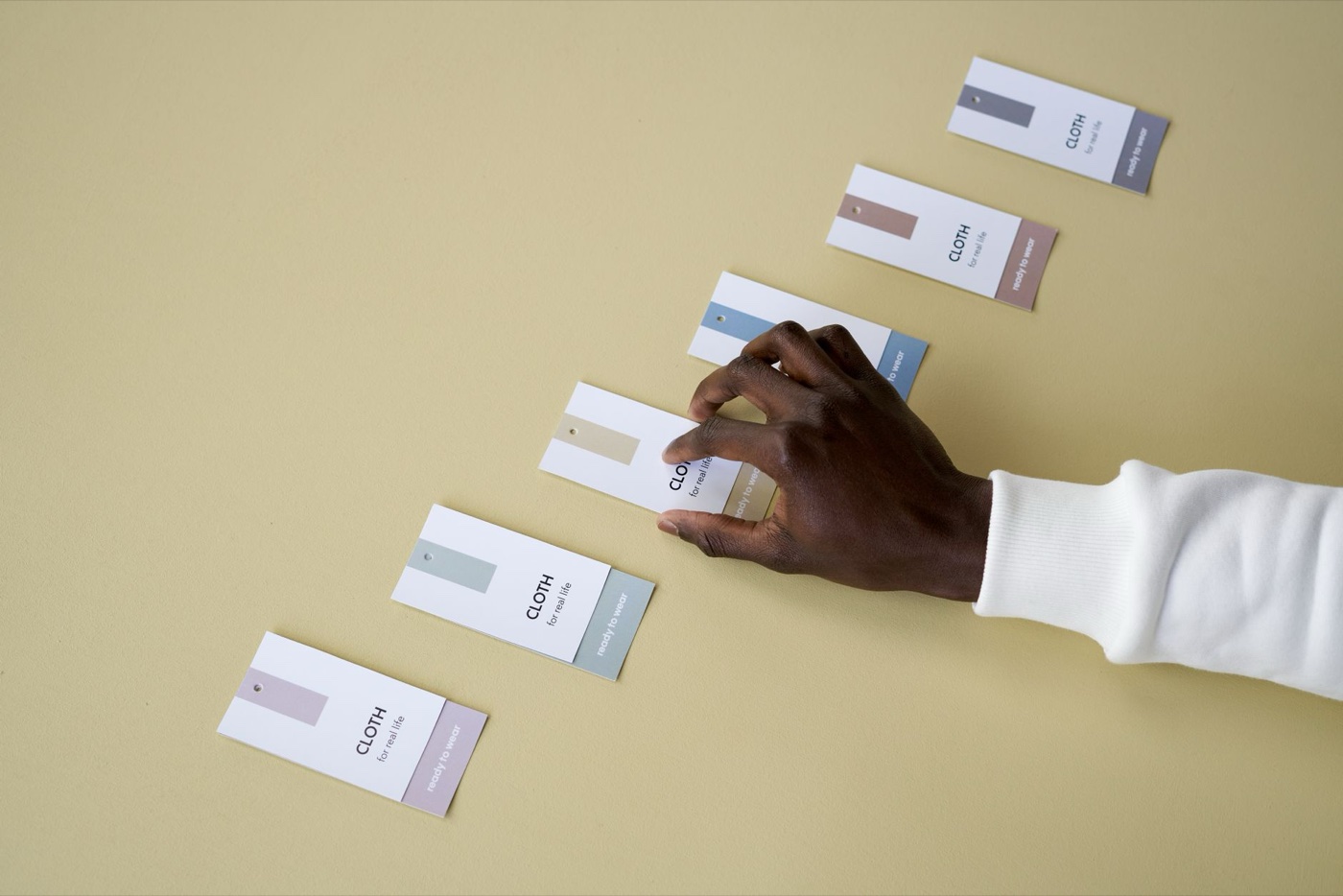 A hand arranging six paper garment tags in a diagonal line on a pale yellow surface, each tag carrying a different colour block