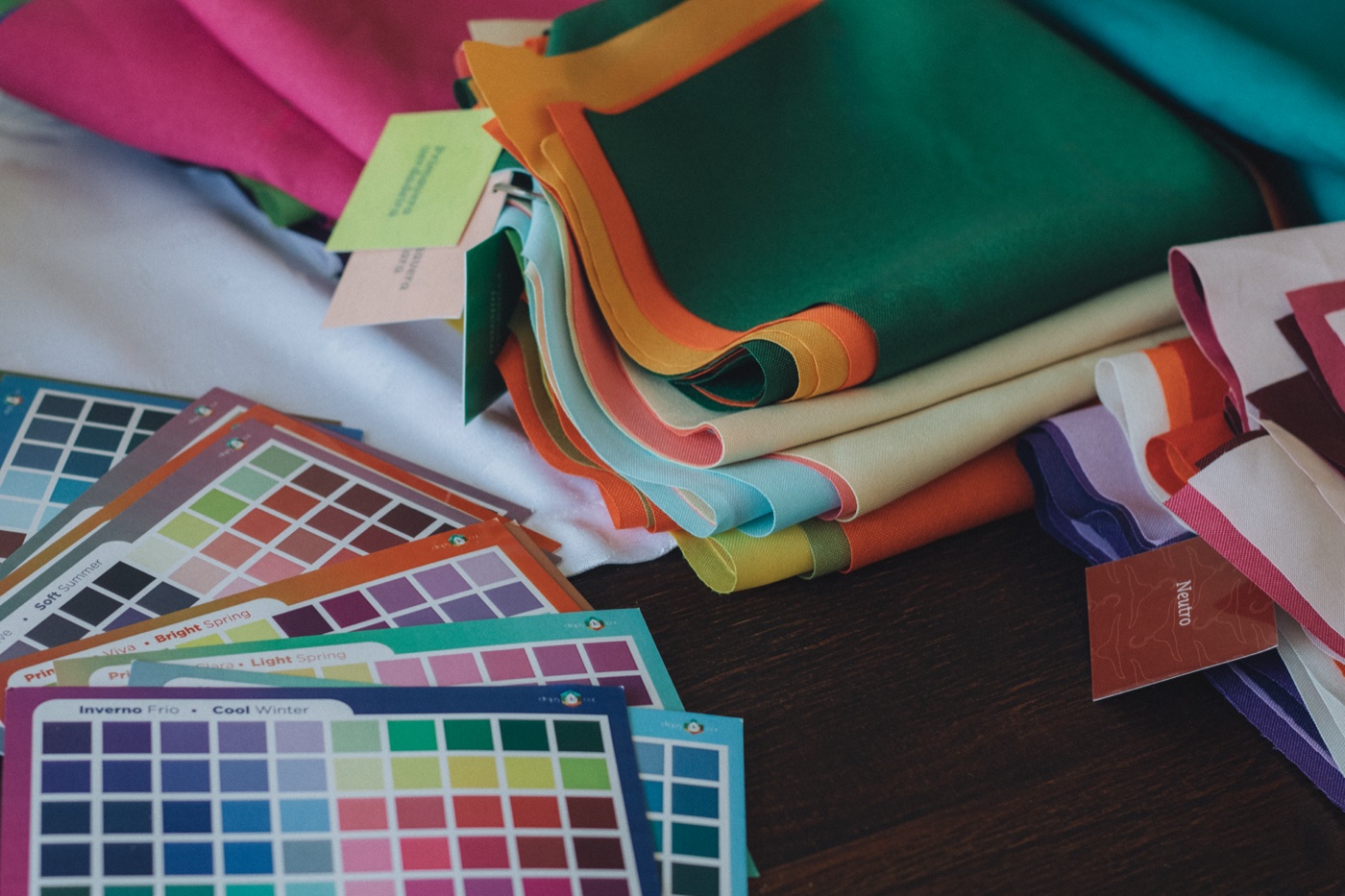 Folded fabric lengths in green, cream, orange and blue stacked beside printed colour palette cards on a dark wooden table