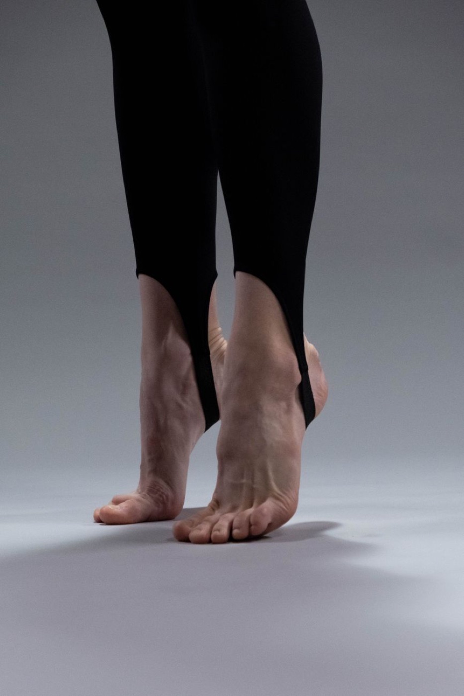 Close-up of the lower legs and bare feet of a person wearing black stirrup leggings on a grey studio floor