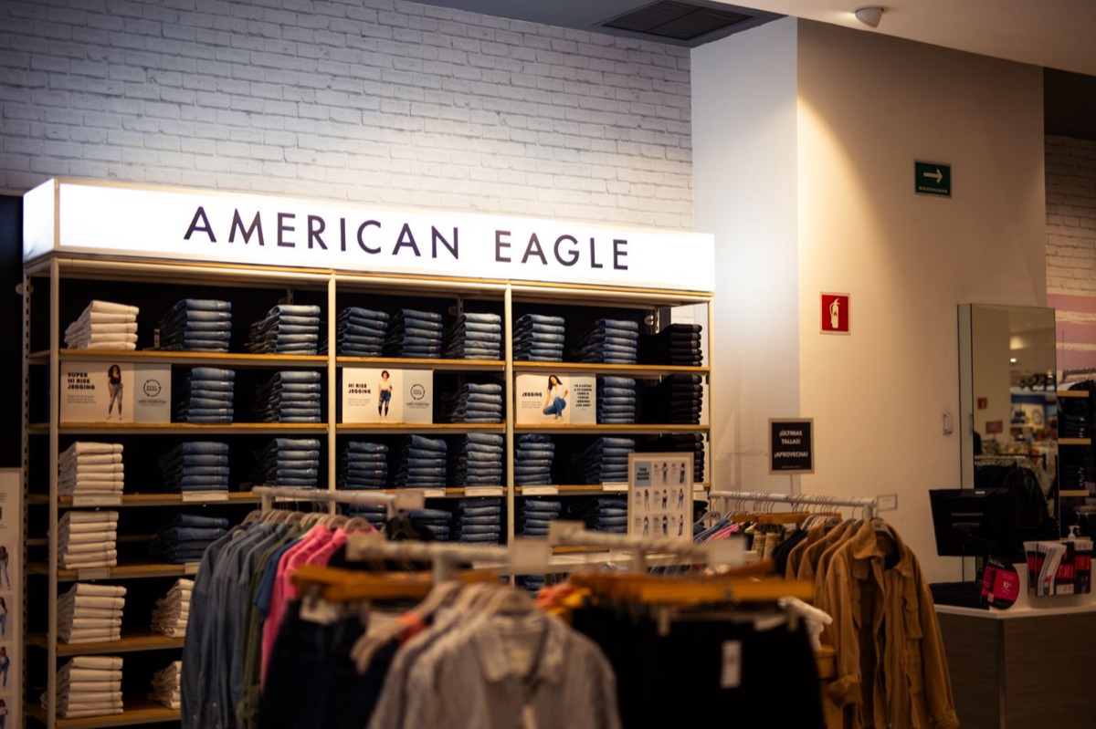 An American Eagle in-store denim wall, folded jeans stacked by wash on white shelves under a lit sign, racks of shirts in front