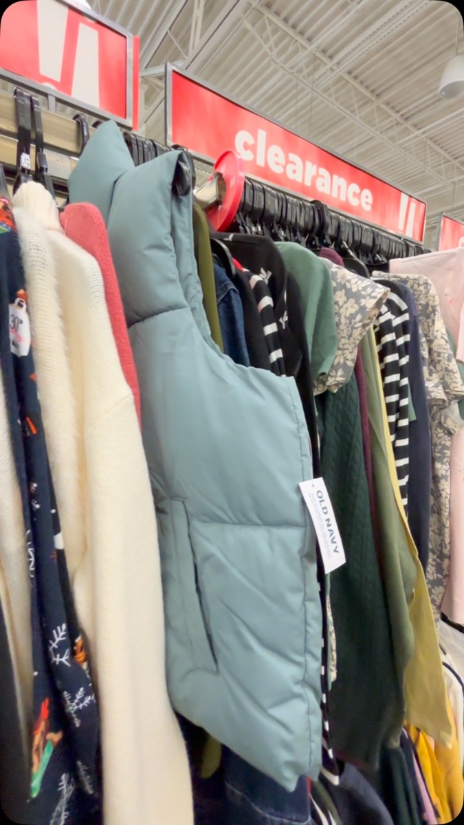 A crowded clearance rack under a red clearance sign, a sage puffer vest with its paper tag still on hanging in front of fleece and floral pieces