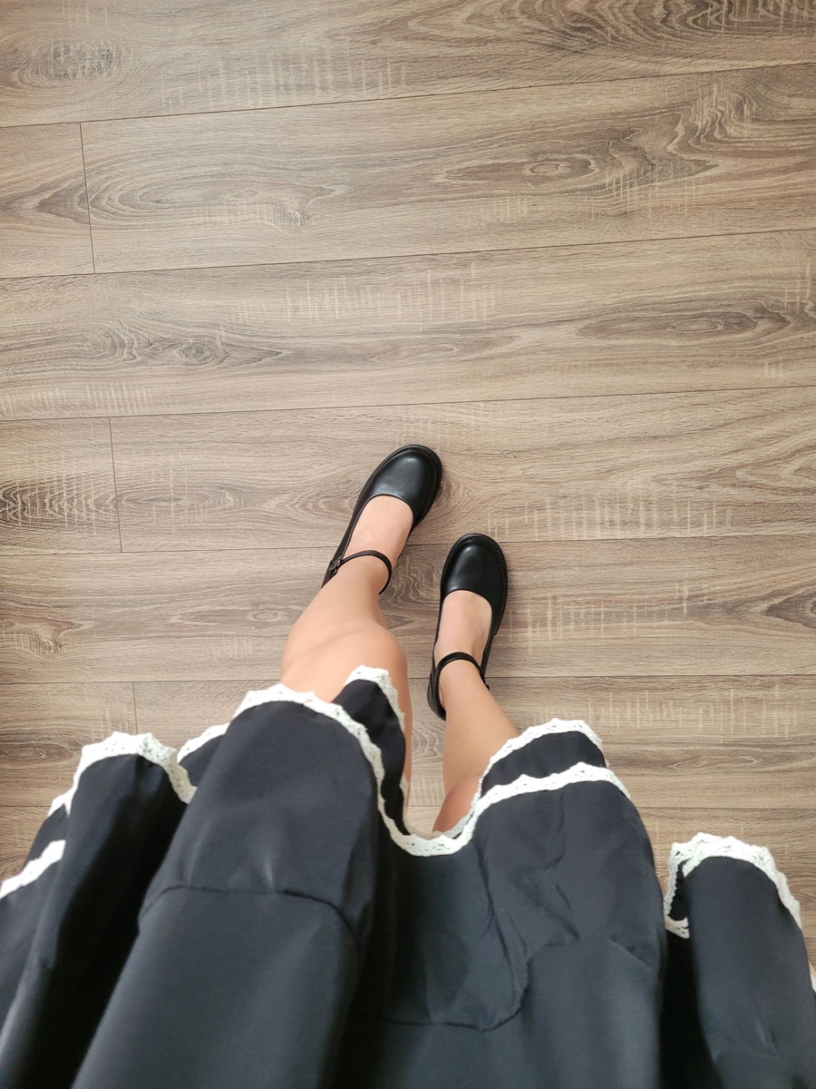 Looking down at black ankle-strap flats worn with a full black skirt trimmed in white lace, on a wood floor