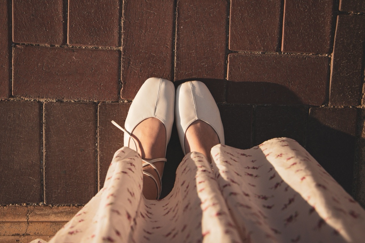 White ballet flats with slim ankle ties worn with a floral dress, seen from above on a brick path in low sun