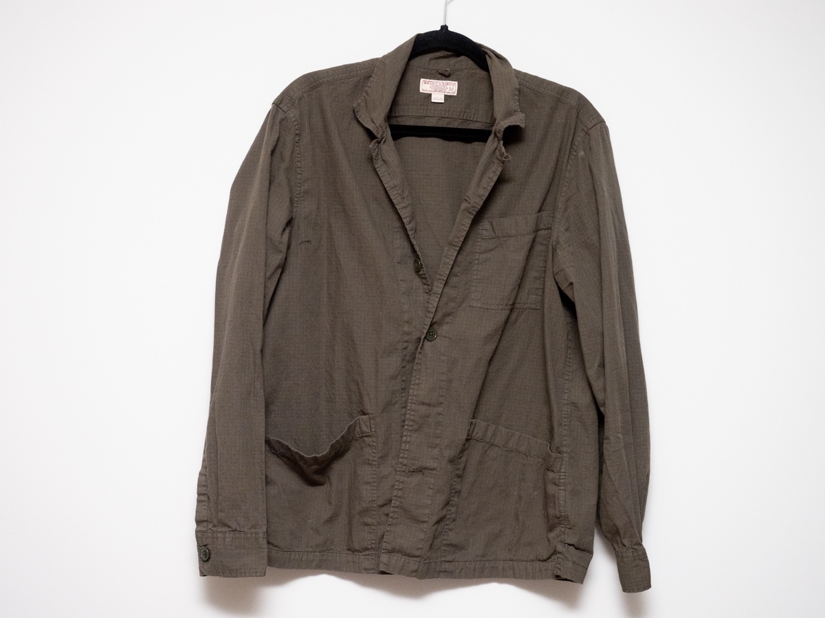 A worn olive cotton chore jacket on a black hanger against a white wall, plain collar and patch pockets