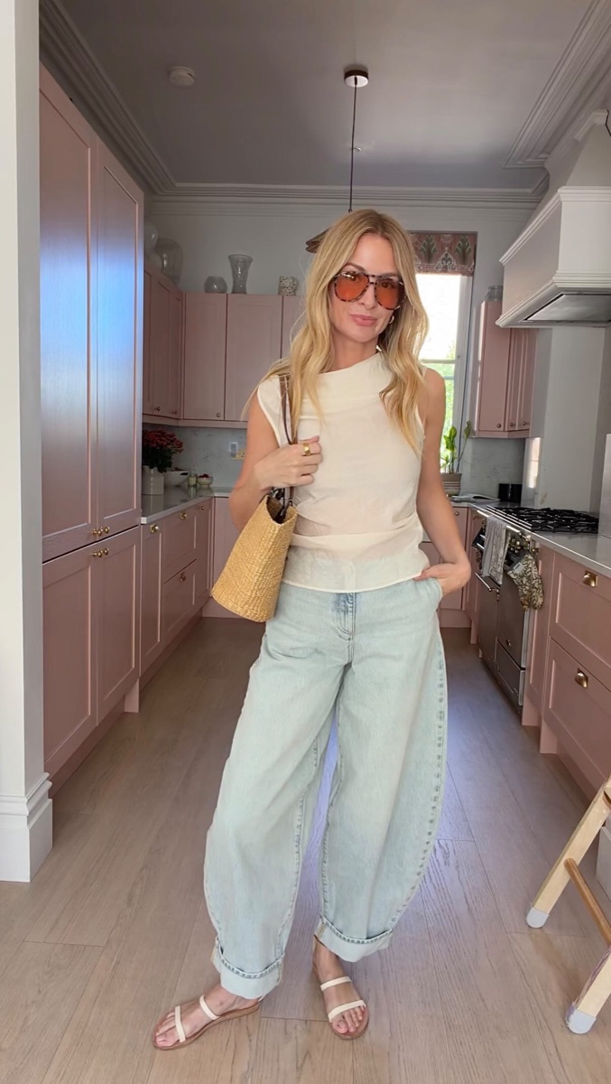 A woman in a pink kitchen wearing pale-wash cuffed barrel jeans with a cream sleeveless top and flat sandals, one hand in her pocket
