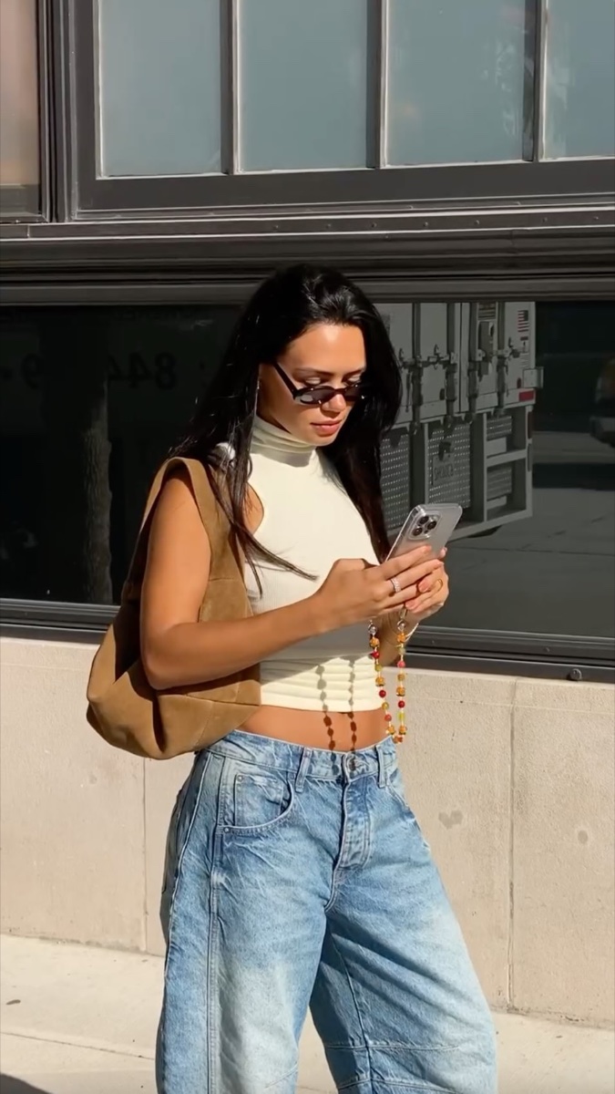 A woman on a New York sidewalk in light-wash barrel jeans, a cream turtleneck crop top, and a suede shoulder bag, checking her phone in hard sunlight