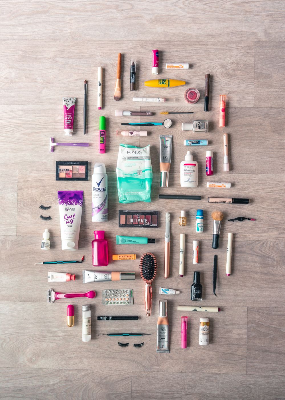 Dozens of makeup and skincare products arranged in an oval on a pale wood floor, seen from above