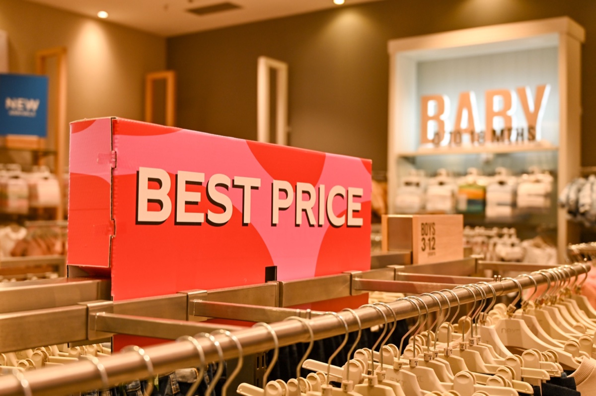 A red and pink cardboard sign reading BEST PRICE standing on a metal rail above a long rack of clothes on hangers