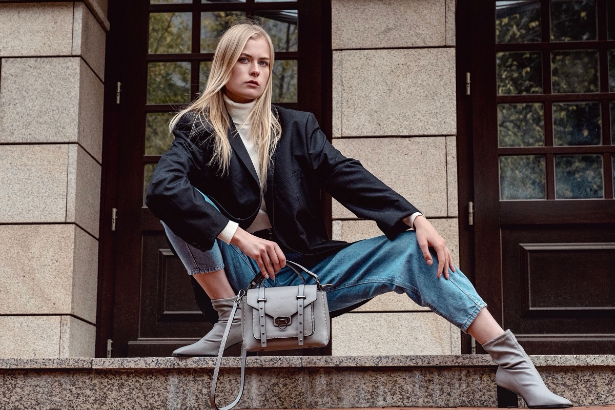 A woman sits on granite steps in an oversized black blazer over a white turtleneck, light-wash jeans and gray ankle boots
