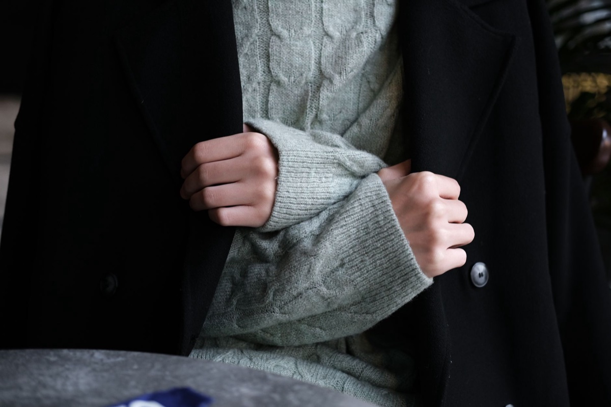 Close view of a black wool coat worn open over a pale grey cable-knit sweater, hands folding back one knit cuff