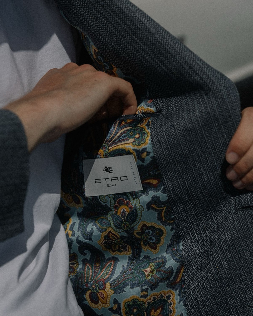 Hands opening a gray herringbone blazer to show a paisley lining and the brand label stitched inside