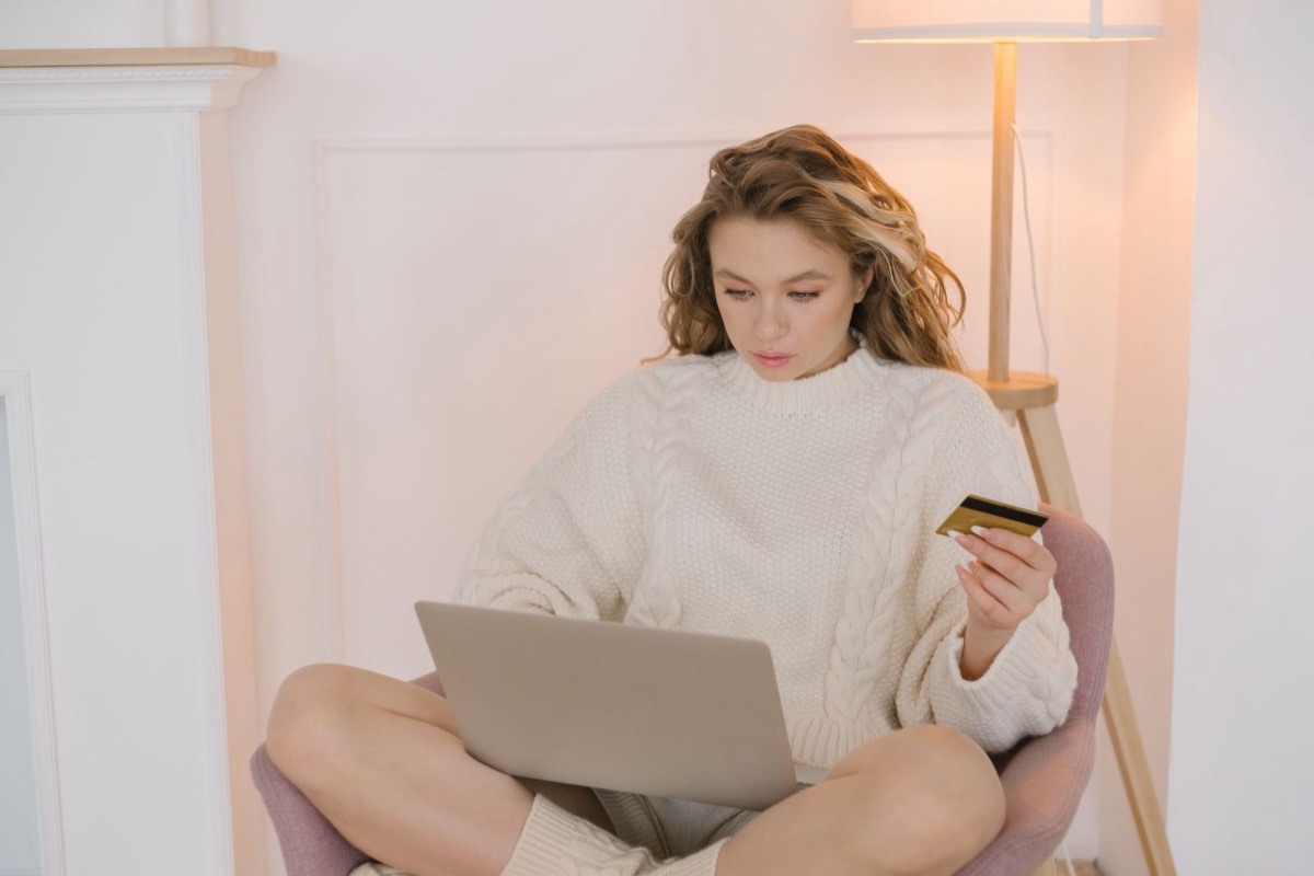 A woman in a cream cable-knit sweater sitting cross-legged with a laptop, holding a gold payment card