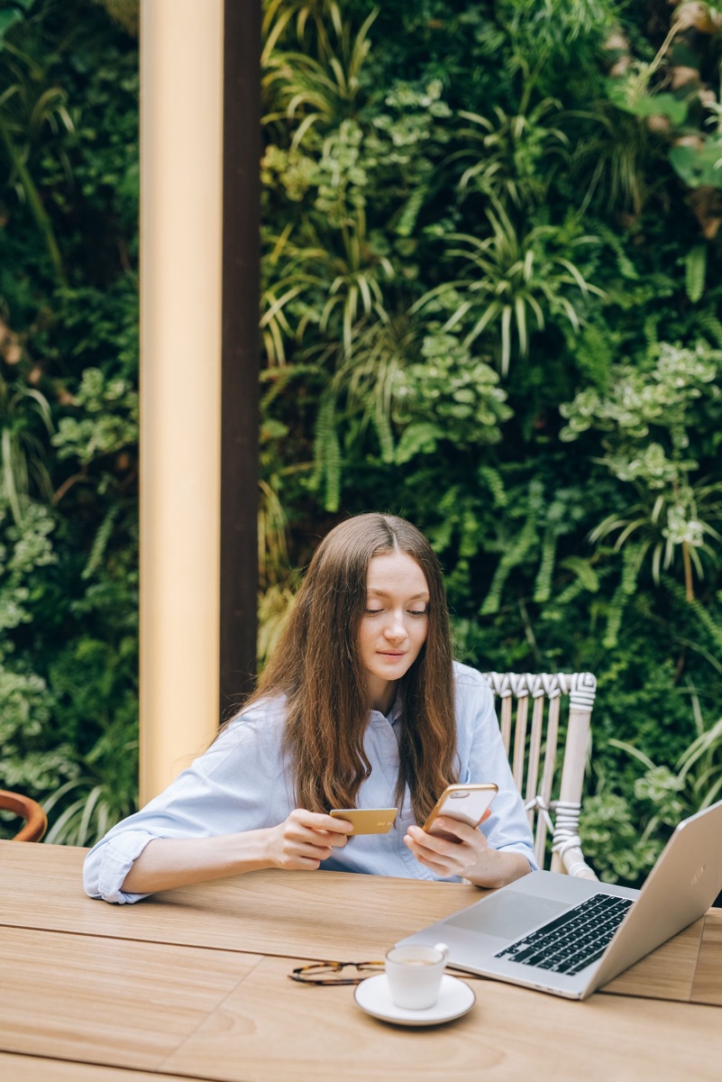 A woman at an outdoor table holding a phone in one hand and a payment card in the other, laptop open in front of her