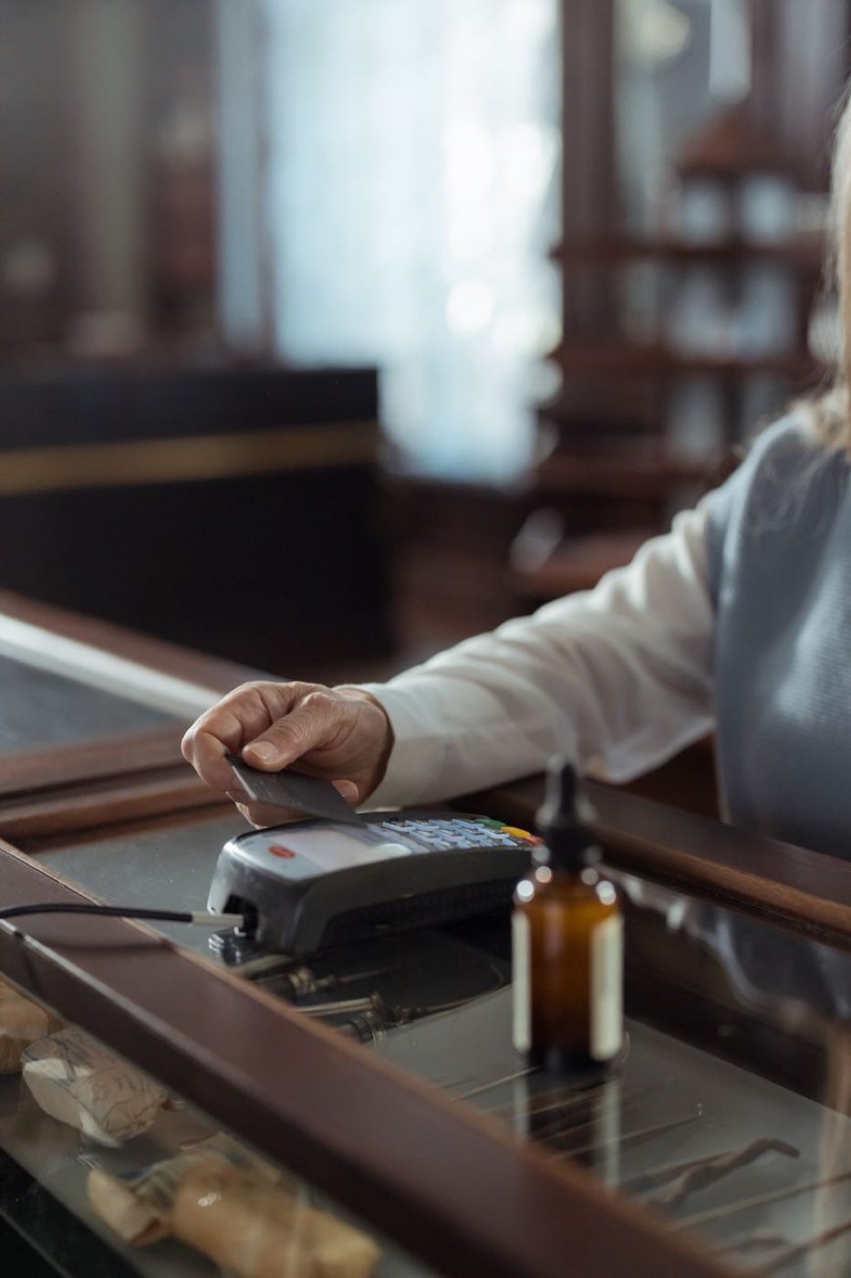 A hand tapping a dark payment card against a card terminal on a polished wooden shop counter