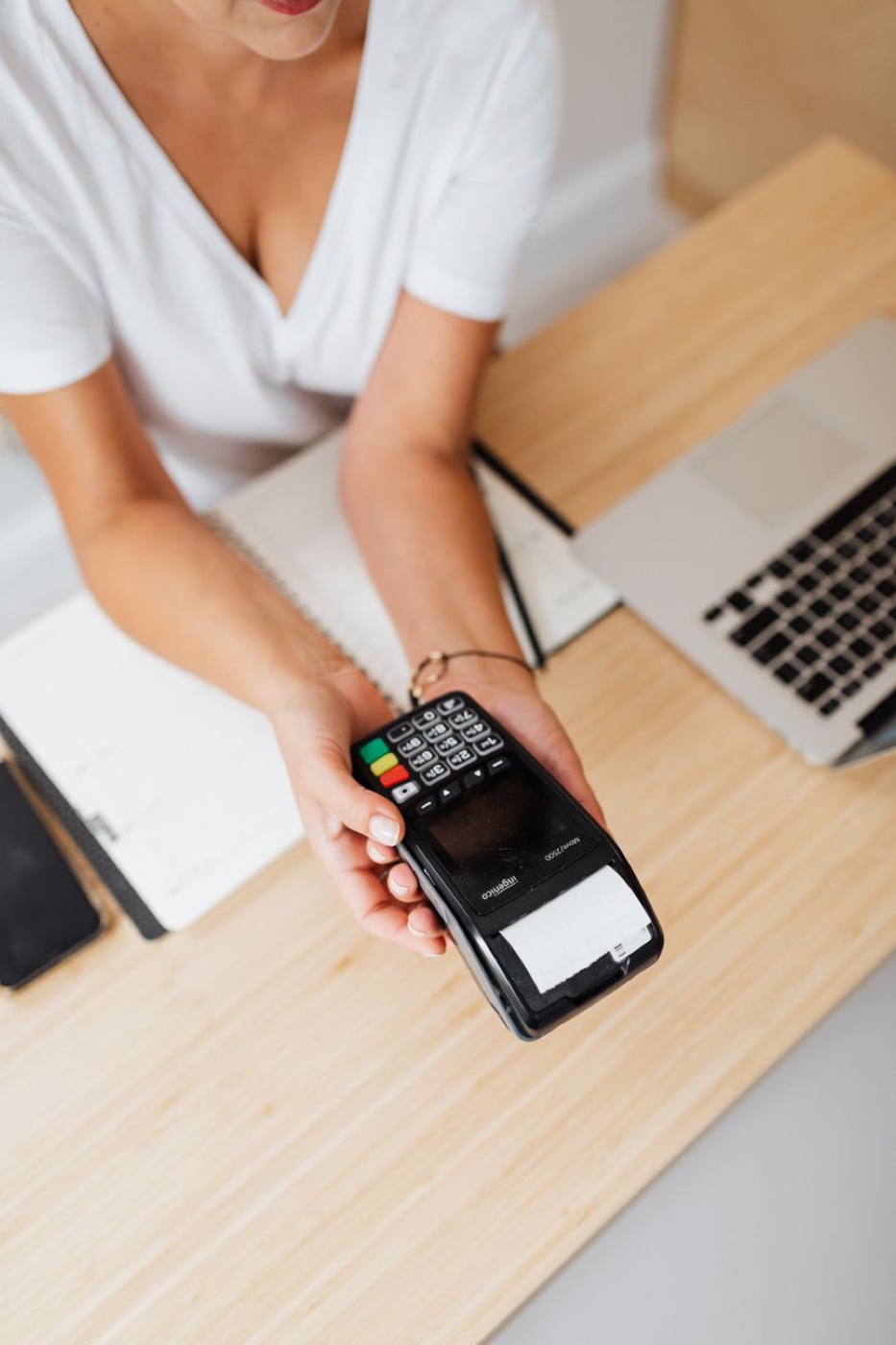 A hand holding a card terminal with a printed receipt curling from the slot, seen from above over a pale wooden desk