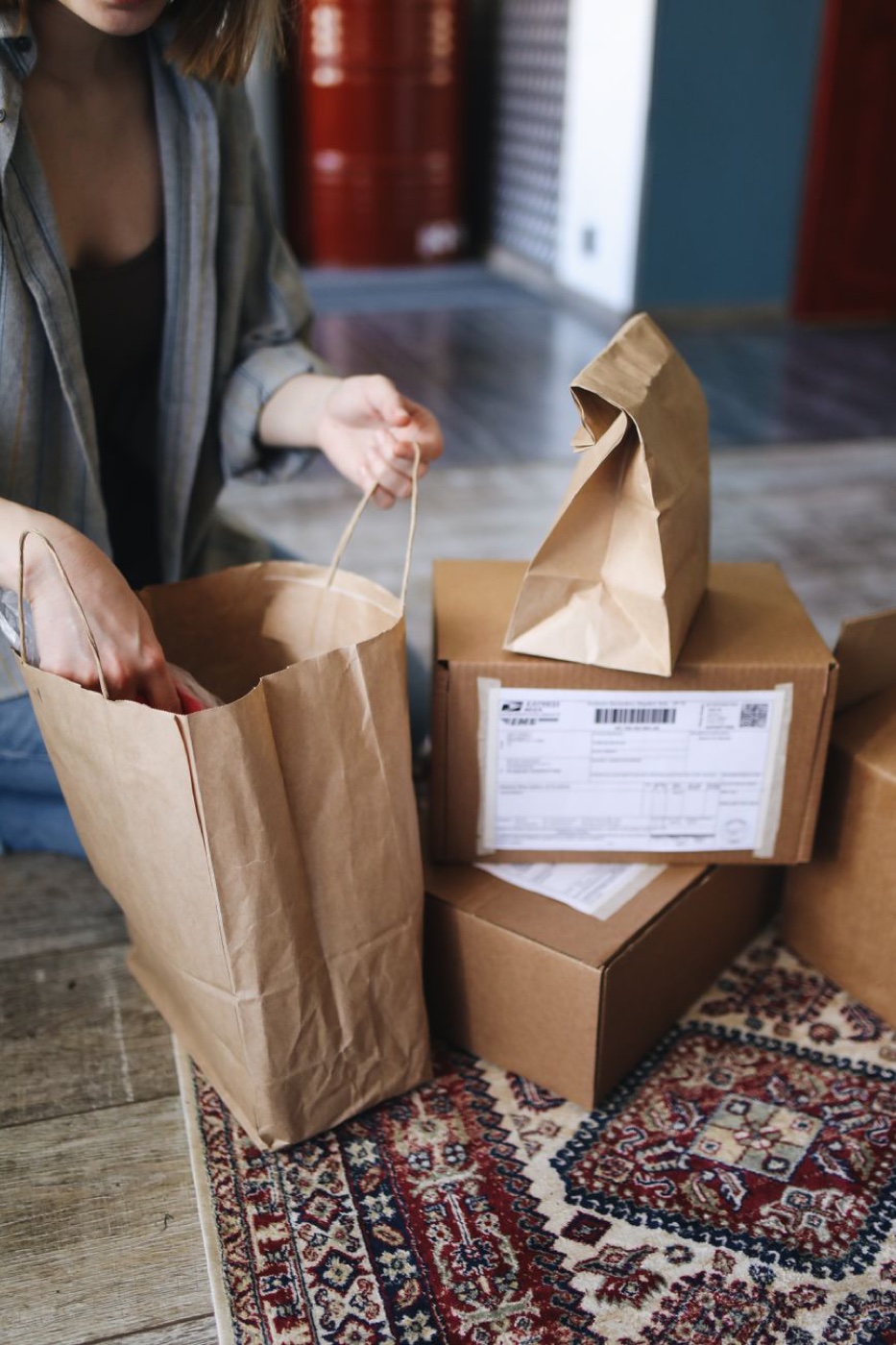 Two cardboard shipping boxes with USPS Express Mail labels stacked beside paper shopping bags on a patterned rug