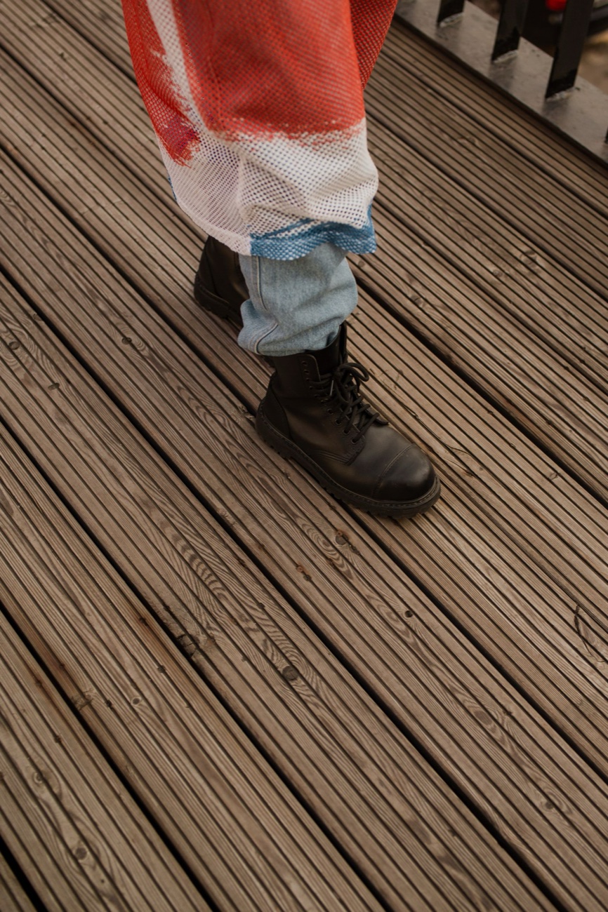 Black leather lace-up combat boots worn with light jeans and a red mesh top, standing on ridged wooden decking
