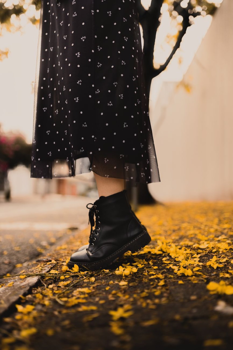 A black lace-up ankle boot below a polka-dot skirt, standing on a pavement covered in fallen yellow blossom