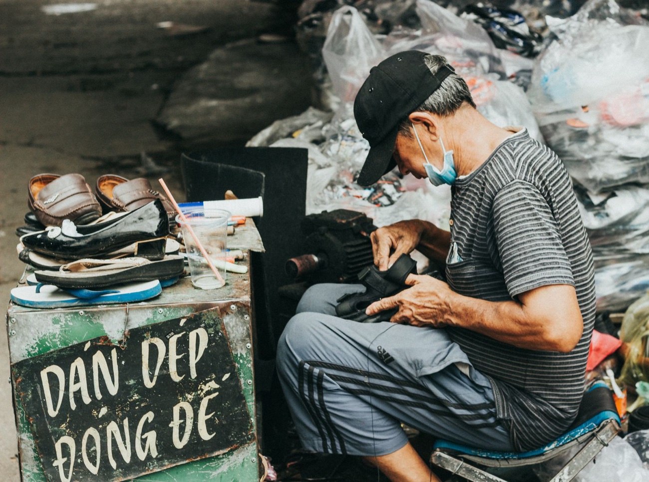 A street cobbler in a cap and face mask stitching a shoe at a sidewalk stand stacked with worn footwear