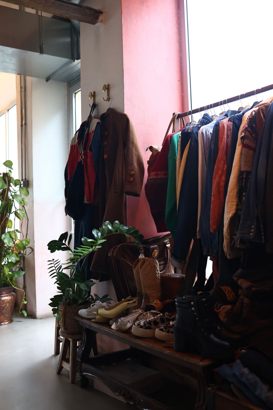 A secondhand clothing shop interior with coats hung on wall hooks, a long rack of jackets, and shoes lined up on a wooden bench in daylight