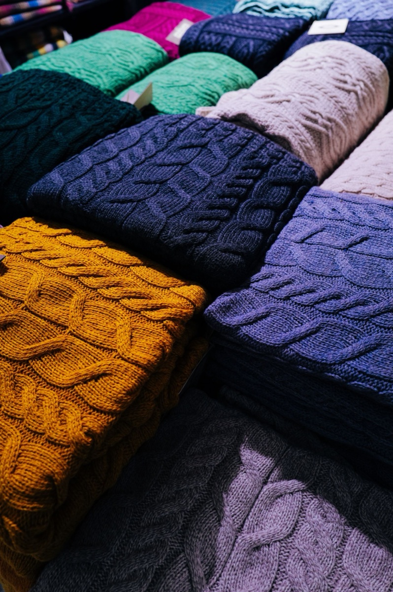 Stacks of folded cable-knit sweaters in mustard, navy, purple and green with paper tags tucked between them on a display table