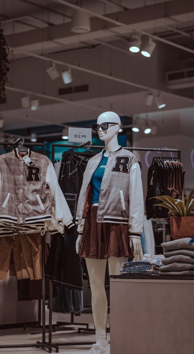 A mannequin in a plaid varsity jacket and brown pleated skirt in a store, with a $12.99 price sign over the racks behind it