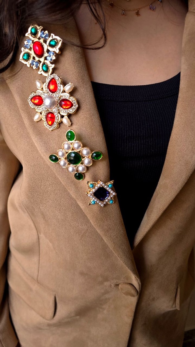 Four jeweled brooches, red and green glass stones set in gold with pearls, pinned in a column down the lapel of a camel suede blazer over a black top
