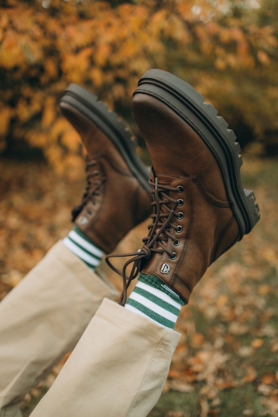 A pair of brown lace-up boots with lug soles worn over green striped socks, feet raised against a backdrop of orange autumn leaves