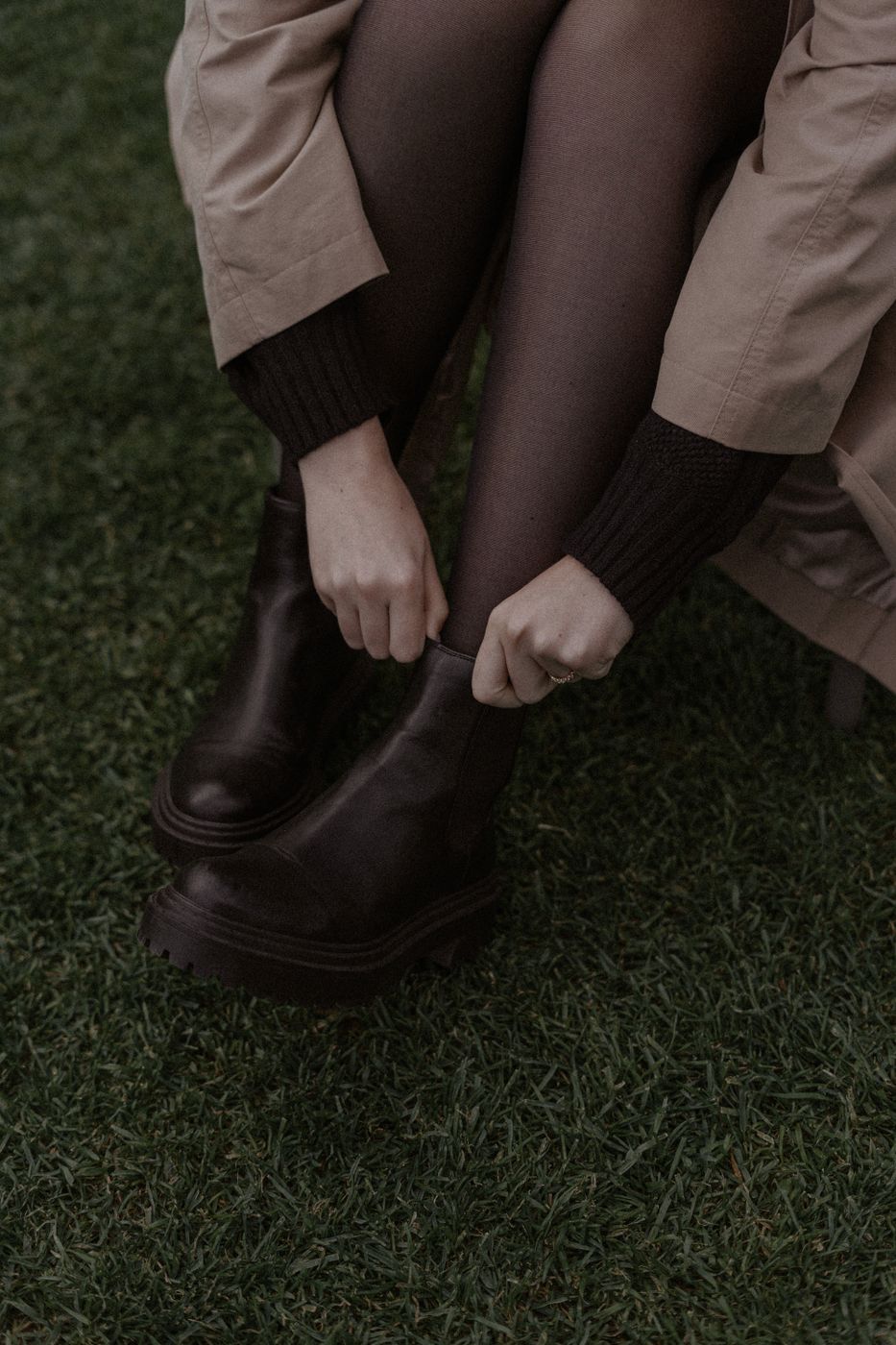 A seated woman in brown opaque tights and a trench coat pulling on a chunky brown boot outdoors