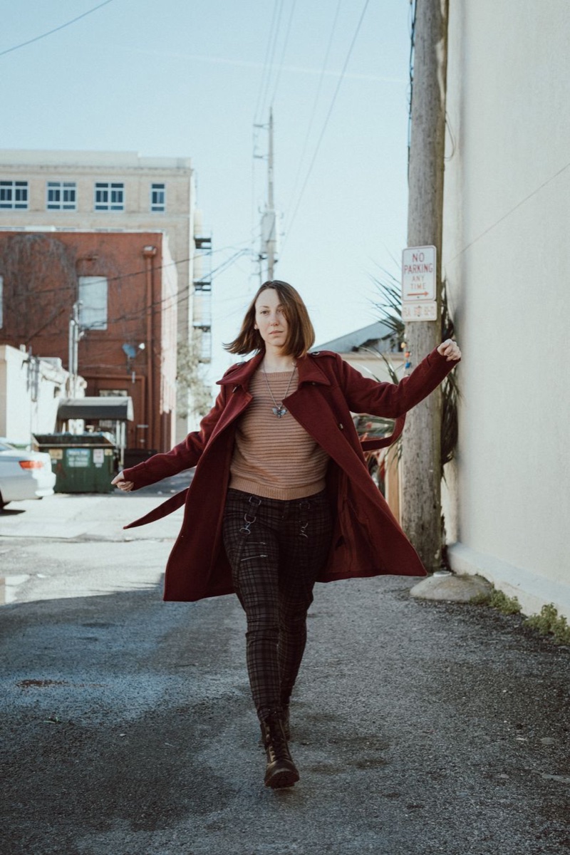 A woman walks through a sunlit alley in a long burgundy trench coat over a tan sweater and plaid trousers