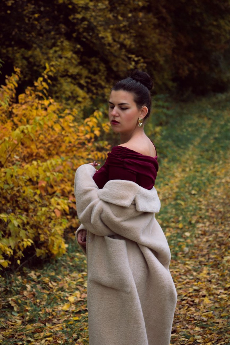 A woman in a dark burgundy off-shoulder top drapes a pale faux fur coat over her arms on a leaf-covered woodland path