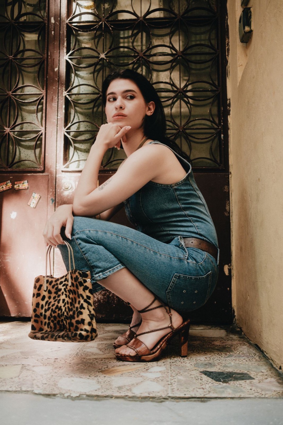 Woman in a denim jumpsuit crouching in a doorway, a small leopard-spotted hair-textured tote in one hand