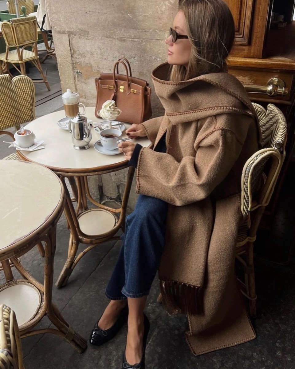A woman at a marble cafe table in a camel scarf coat with the attached scarf wrapped high around her neck, coffee service and a brown leather bag beside her