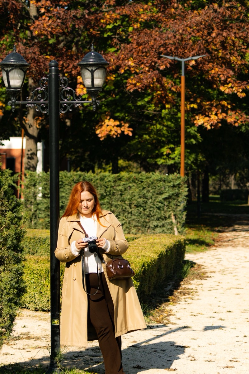 A woman in a camel trench coat and dark brown trousers checking a camera in a park with early fall foliage