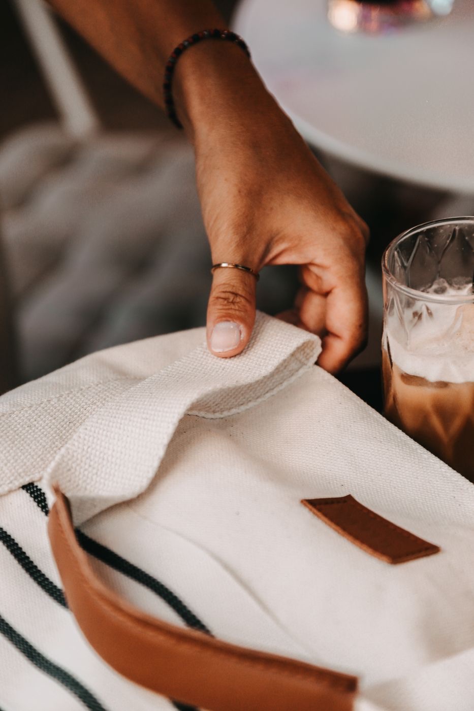 A hand pinching the woven strap of a cream canvas tote with tan trim at a cafe table beside an iced coffee