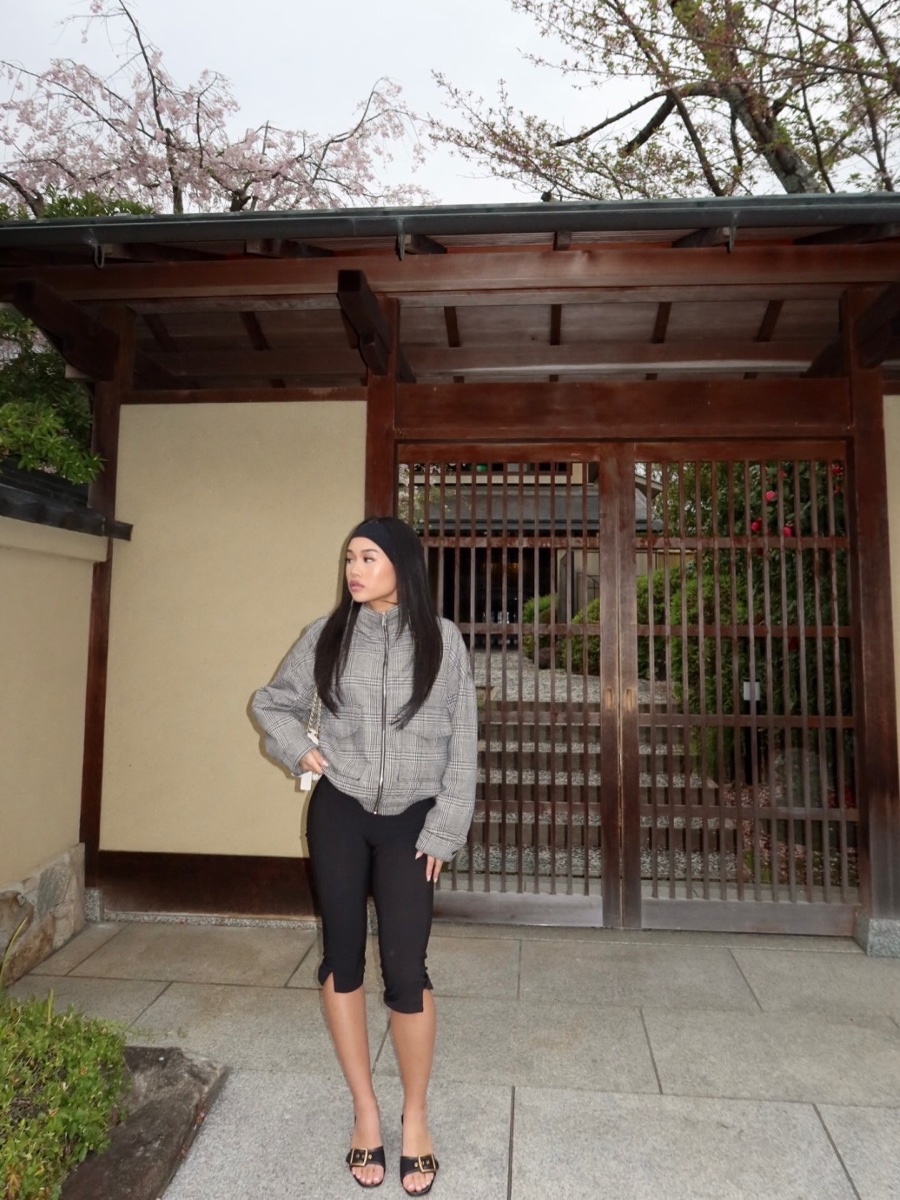 A woman in black capri leggings, a grey plaid zip jacket and buckled black sandals stands in front of a wooden gate