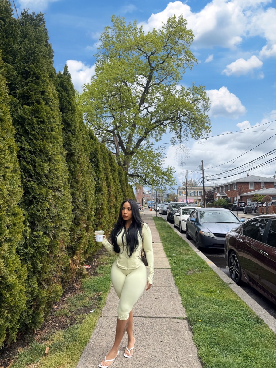 A woman stands on a suburban sidewalk in a matching pale green capri legging set with white flip-flops, holding an iced drink