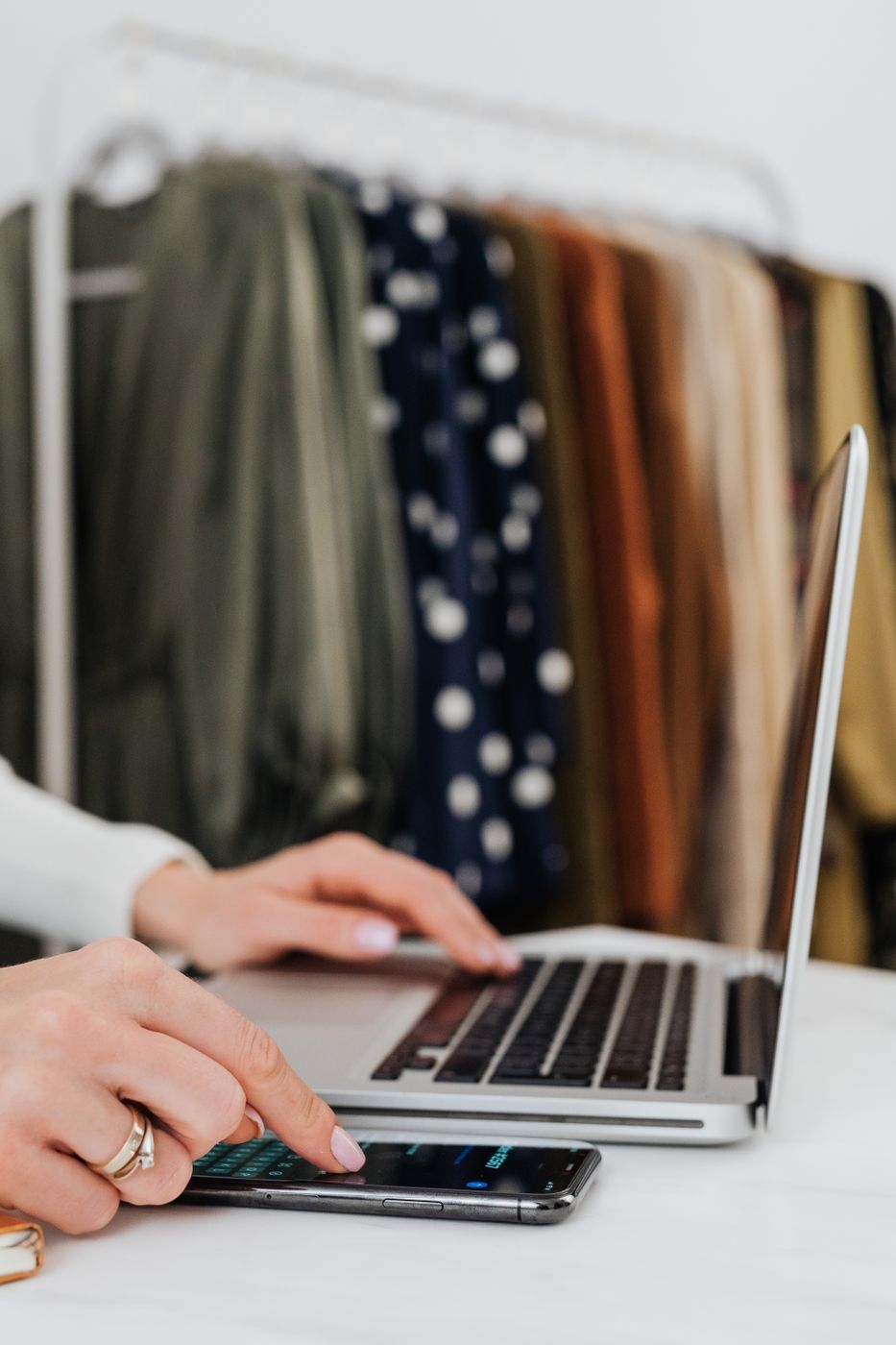A hand tapping a smartphone beside an open laptop on a white desk, with a rack of green, navy and rust clothing blurred in the background