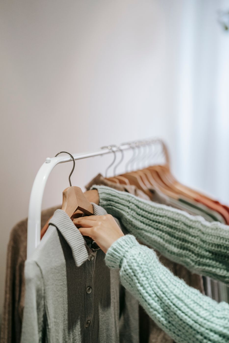 A woman in a mint knit sweater lifting a wooden hanger holding a gray collared cardigan from a white clothing rail, her face out of frame