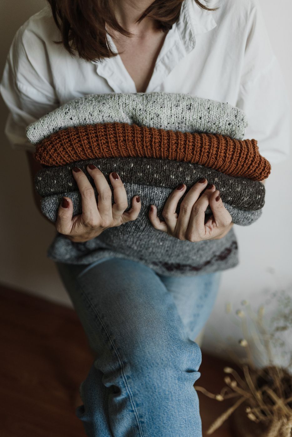 A seated woman in a white shirt and blue jeans holding a folded stack of five knit sweaters in cream, rust and gray, her face out of frame