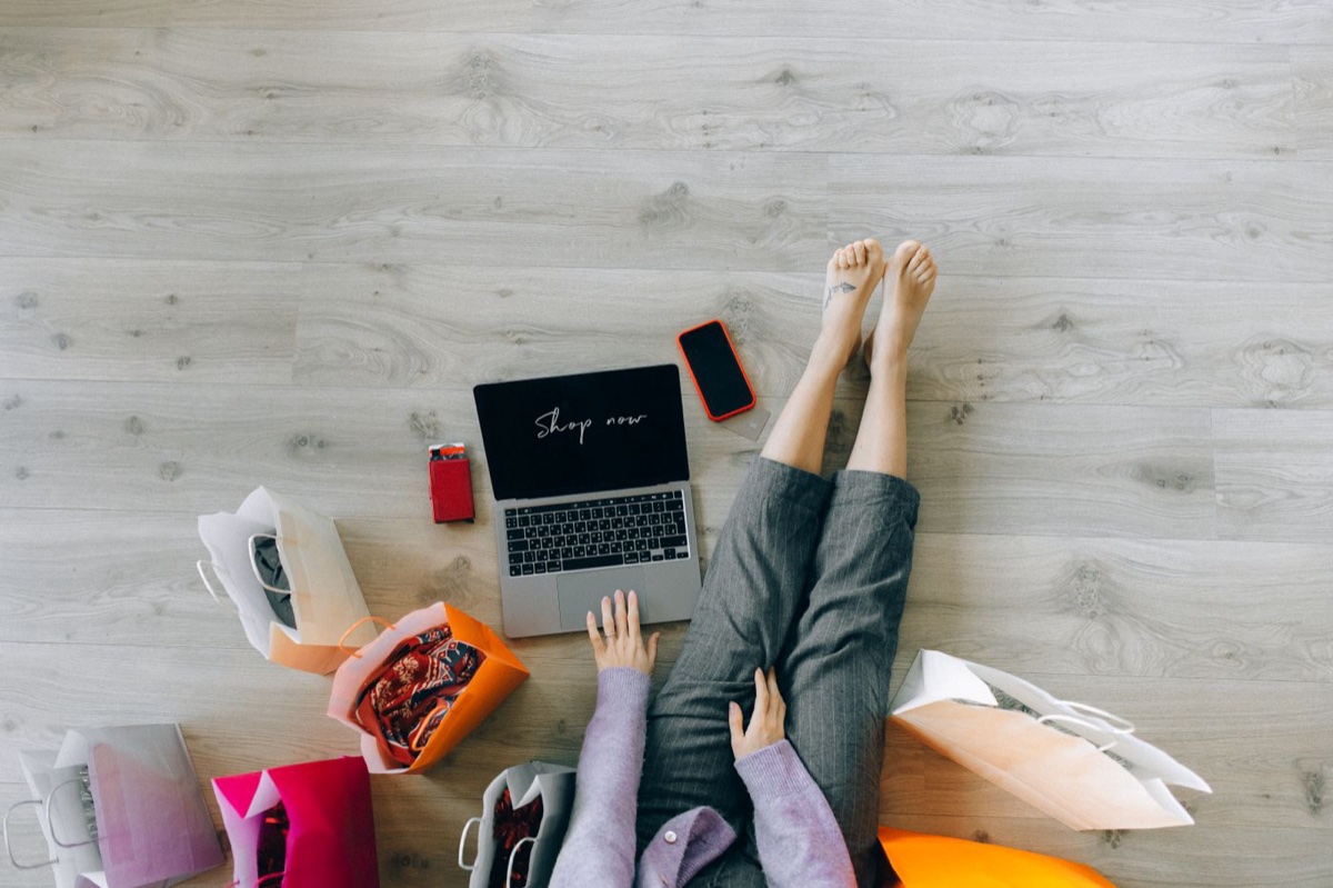 Overhead view of a shopper on a wood floor with a laptop reading Shop now, a phone, a card holder and paper shopping bags