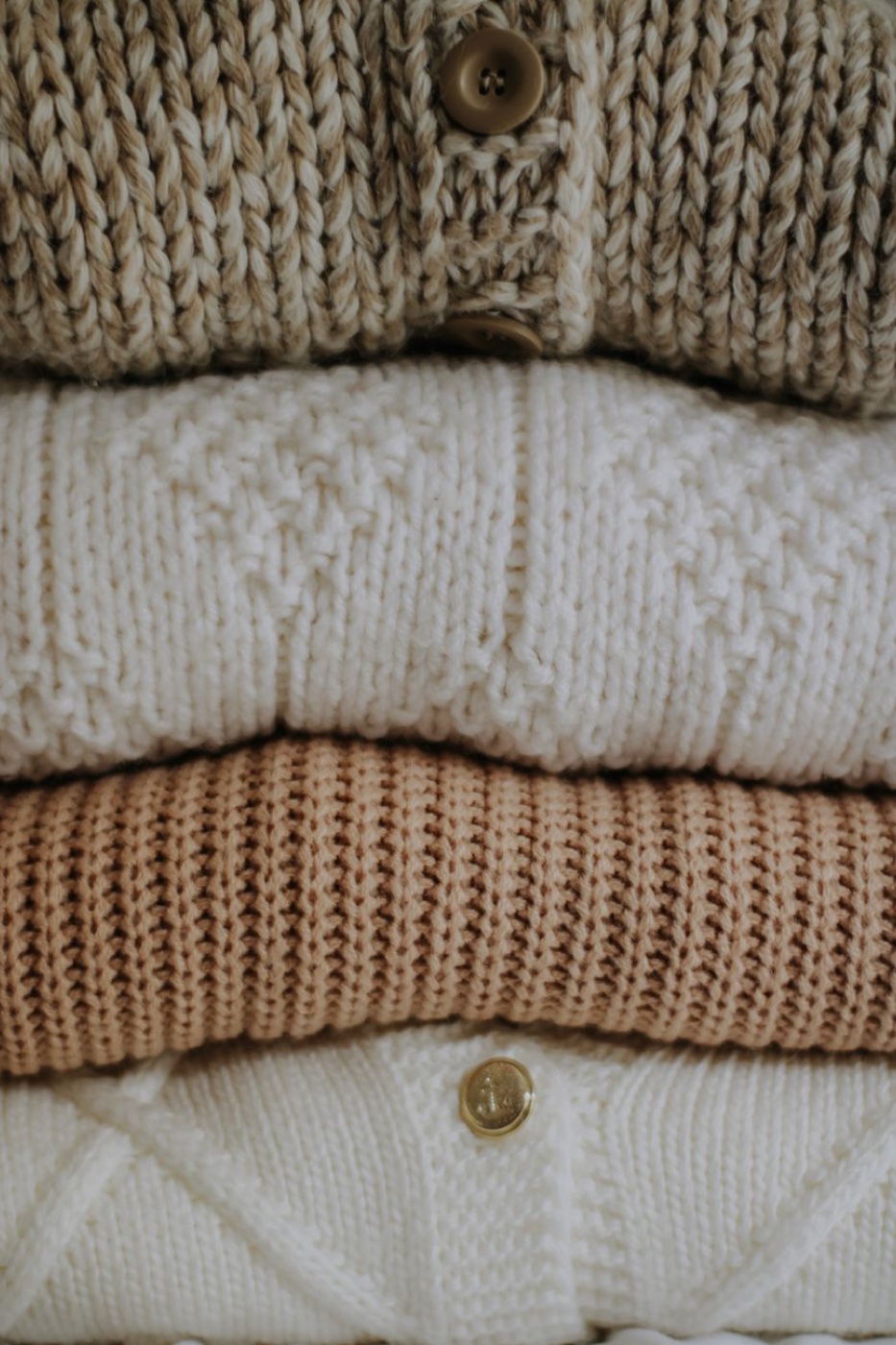 Four folded knits stacked, a chunky oatmeal cardigan with horn buttons on top, then a fine ivory knit, a mid-weight camel rib and a cream cable knit